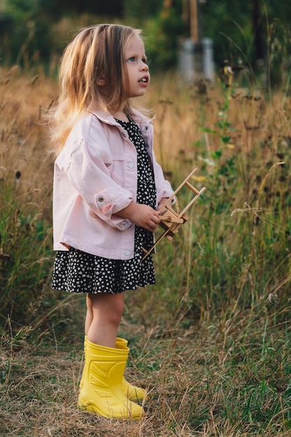 Una bambina sta giocando con un aereo di legno in un villaggio in un campo la felicità dei bambini felice concetto di infanzia una bella ragazza bionda in un vestito e stivali sembra pensieroso