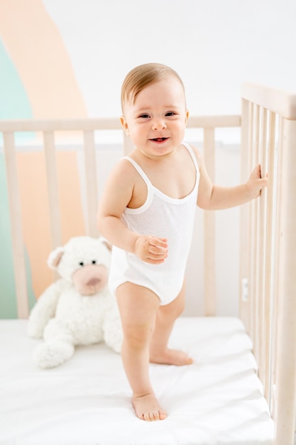 una bambina sta in piedi nella culla e sorride o ride a casa nella nursery in uno stile di vita bianco