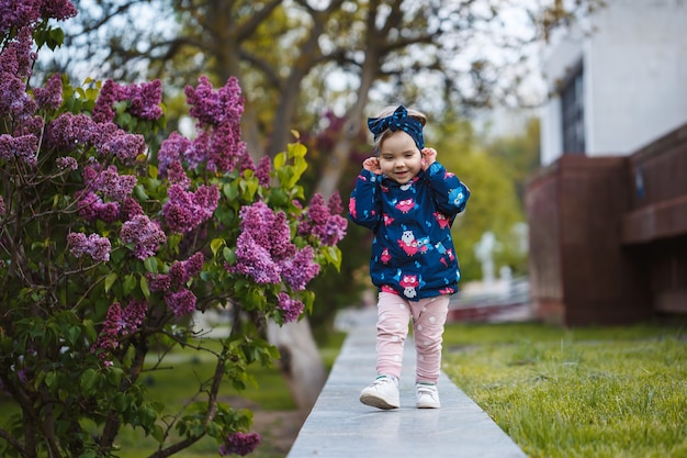 Una bambina sta vicino a un rigoglioso cespuglio di lillà, sorride e annusa fiori viola