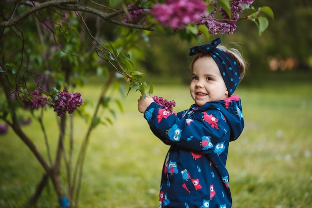 Una bambina sta vicino a un rigoglioso cespuglio di lillà, sorride e annusa fiori viola