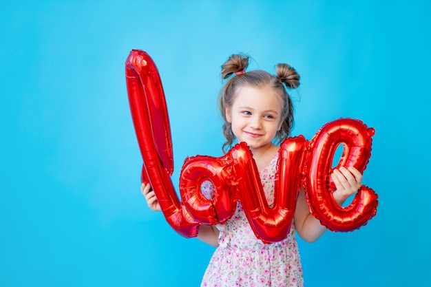 Una bambina su uno sfondo blu isolato per studio con palloncini rossi nella scritta amore si diverte e scherza Spazio per copiare il testo Concetto di San Valentino