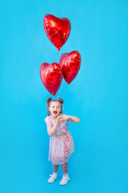 Una bambina su uno sfondo blu per studio isolato con palloncini rossi a forma di cuore si sta divertendo e scherzando Spazio per copiare il testo Concetto di San Valentino