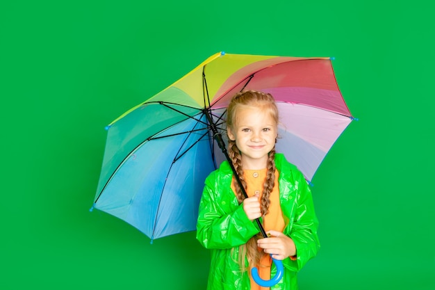 Una bambina su uno sfondo verde isolato con un ombrello e un impermeabile sorride, spazio per il testo