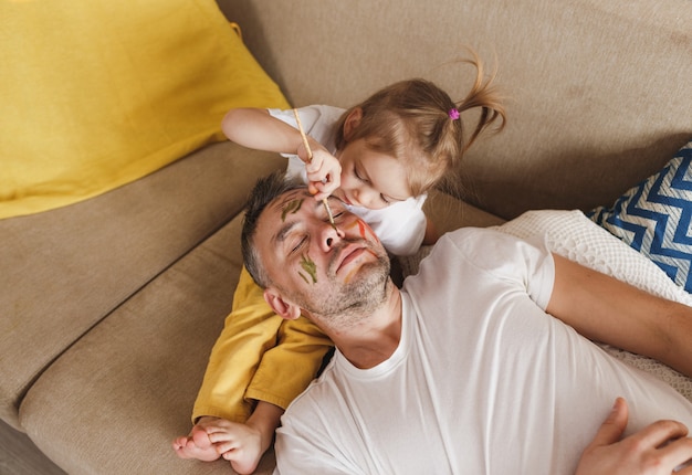 Una bambina sul divano dipinge con cura il viso di suo padre durante i giochi di famiglia insieme