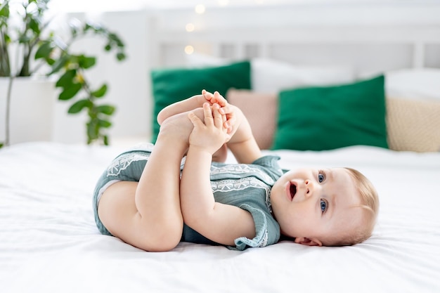 una bambina sul letto a casa in un bodysuit verde giace sulla schiena e gioca con le gambe le leccandole con la bocca sorridendo o ridendo bambino carino divertente a casa prima di andare a letto