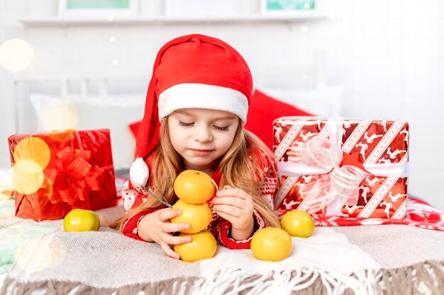 Una bambina sul letto di casa che gioca con i mandarini costruendo un pupazzo di neve al mandarino e aspettando il nuovo anno o il Natale con un cappello rosso di Babbo Natale