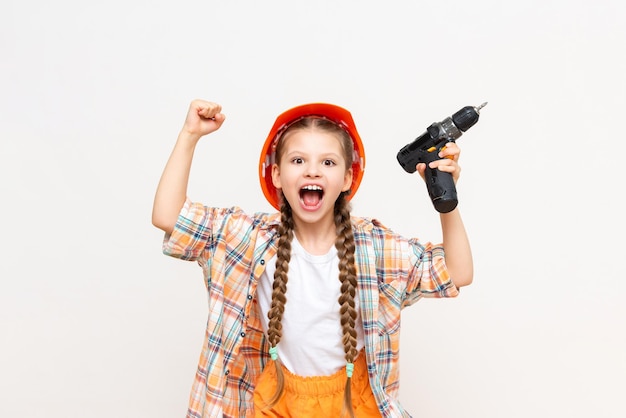 Una bambina tiene un trapano e urla Un bambino con un casco da costruzione su uno sfondo bianco isolato Il concetto di ristrutturazione nella stanza dei bambini