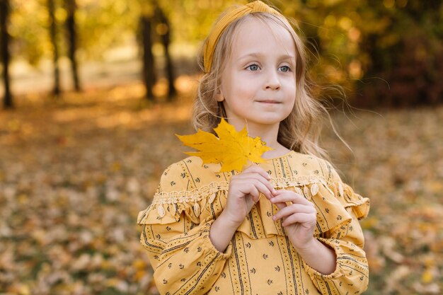 Una bella bambina bionda sta camminando in un parco autunnale con in mano foglie d'acero gialle