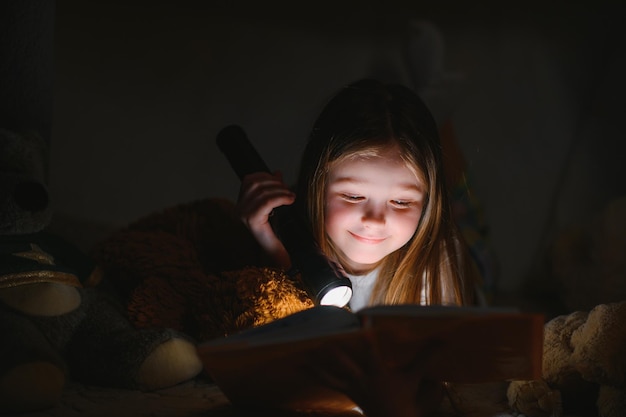 Una bella bambina che legge un libro sul pavimento sotto la lampada. Bambini e istruzione.