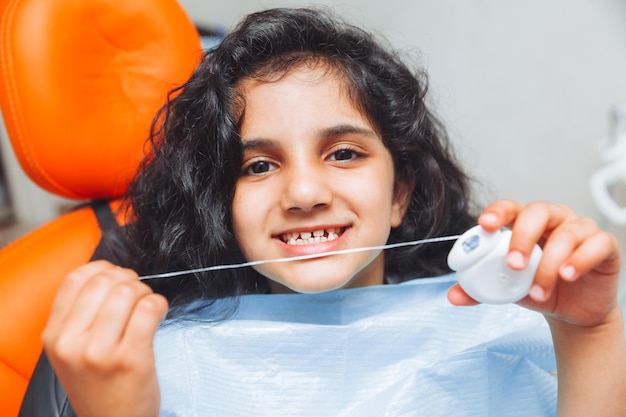 Una bella bambina che usa il filo interdentale su uno sfondo bianco mentre si siede sulla poltrona di un dentista per l'igiene dentale