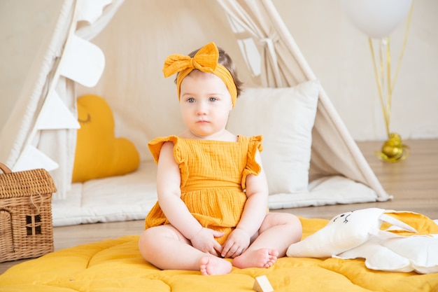 Una bella bambina di un anno in abiti gialli si siede sul pavimento e distoglie lo sguardo