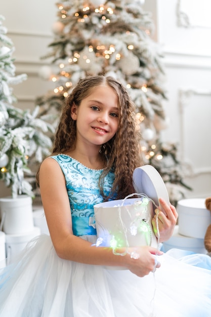 Una bella bambina in un abito festivo si siede vicino all'albero di Natale e tiene i regali