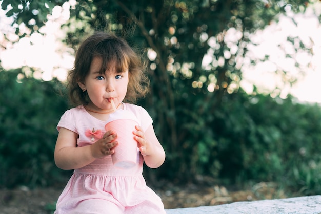 Una bella bambina in un vestito rosa su uno sfondo di piante beve un drink da un bicchiere da cocktail carino