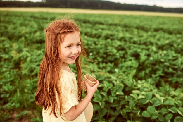 Una bella bambina nel campo verde raccoglie e mangia fragole divertendosi