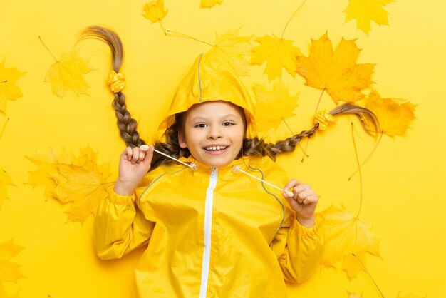 Una bella bambina su uno sfondo giallo autunnale con acero lascia un bambino in un impermeabile impermeabile e un caldo maglione lavorato a maglia