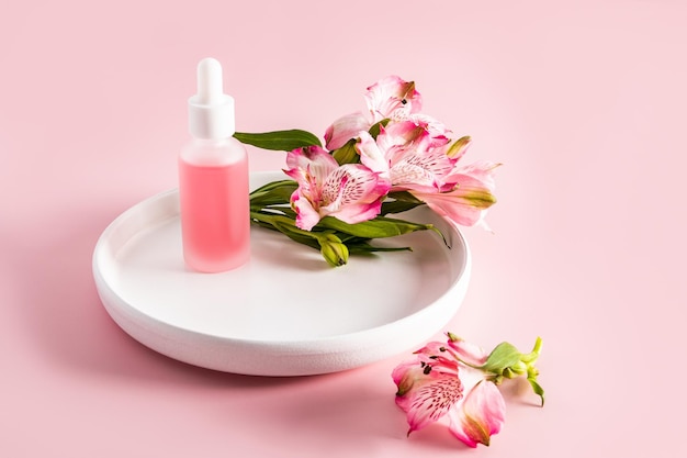 Una bella bottiglia cosmetica con una pipetta bianca si trova su un piatto di ceramica con fiori di astromeria sfondo rosa cosmetici naturali