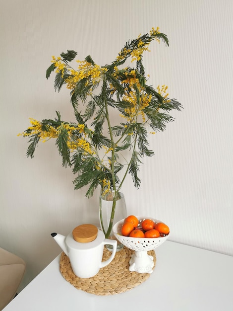 Una bella composizione di un ramo di mimosa un mandarino e una teiera bianca su una tovaglietta di vimini per apparecchiare la tavola Tavolo da cucina bianco e interni