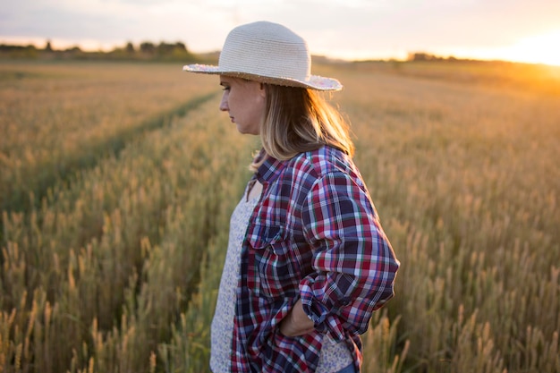 Una bella contadina di mezza età con un cappello di paglia e una camicia a quadri si trova in un campo di grano dorato che matura durante il giorno alla luce del sole