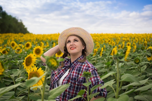 Una bella contadina di mezza età gioiosa e sorridente con un cappello di paglia e una camicia si trova in un campo di girasoli in una giornata di sole