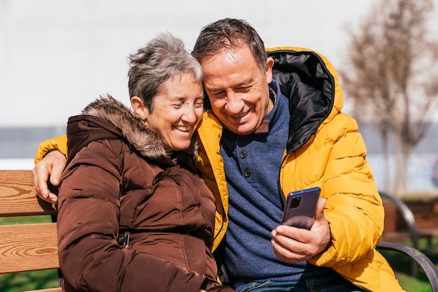 Una bella coppia senior sorridente utilizzando uno smartphone mentre era seduto su una panchina nel parco