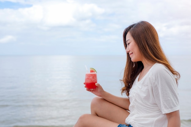 Una bella donna asiatica che beve succo di anguria mentre è seduta in riva al mare