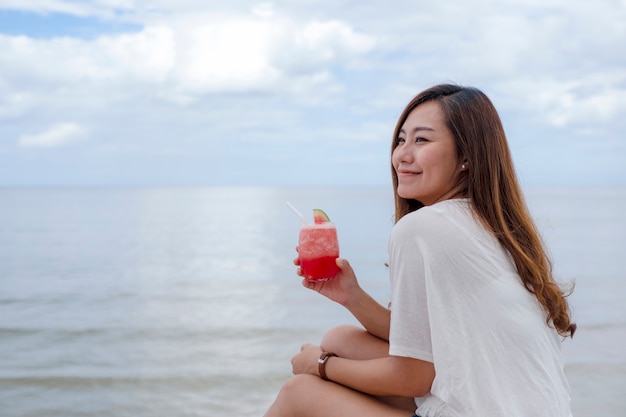 Una bella donna asiatica che beve succo di anguria mentre è seduta in riva al mare