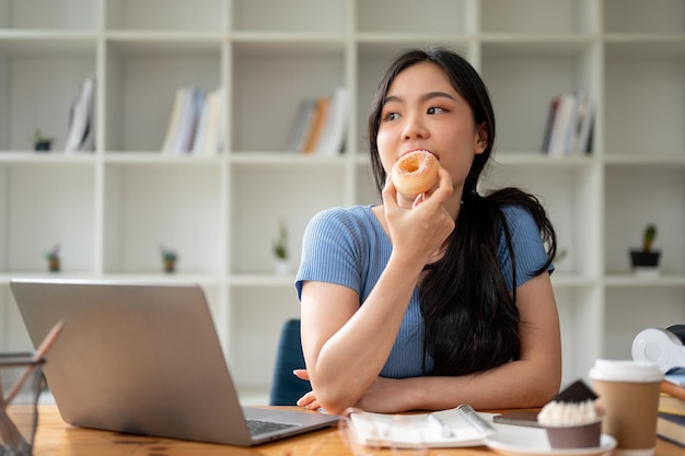 Una bella donna asiatica che si gode il suo spuntino mangiando ciambelle al suo tavolo da studio a casa