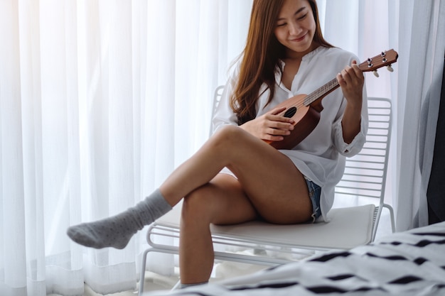 Una bella donna asiatica che si siede e che gioca ukulele in camera da letto