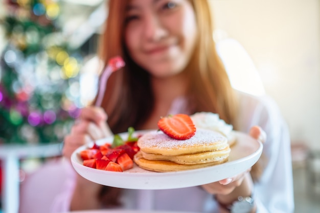 Una bella donna asiatica che tiene e mangia frittelle con fragole e panna montata con la forchetta