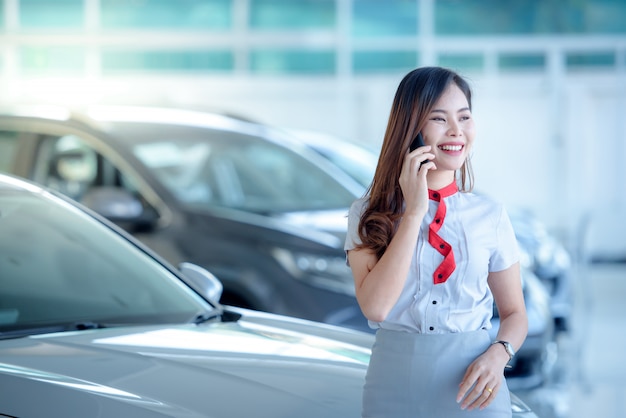 Una bella donna asiatica è felice di vendere una nuova auto nello showroom e divertirsi a parlare al telefono. Entusiasta delle buone notizie online nello showroom.