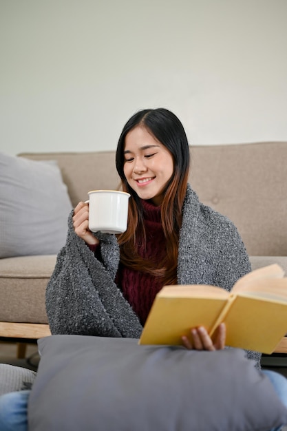 Una bella donna asiatica in un maglione accogliente sta sorseggiando cioccolato caldo mentre legge un libro