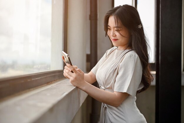 Una bella donna asiatica usa il suo smartphone mentre si trova accanto alla finestra di una torre