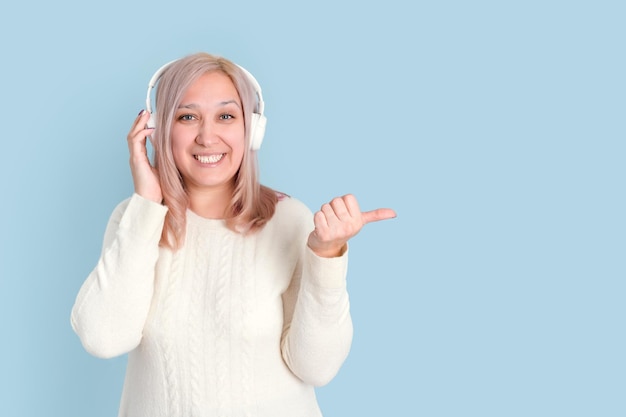Una bella donna bionda di 35-40 anni ascolta musica in cuffie wireless e fa un gesto con il dito in alto mentre si trova su uno sfondo blu.