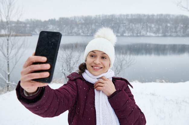 Una bella donna castana che fa un selfie sullo sfondo del paesaggio innevato mentre cade la neve