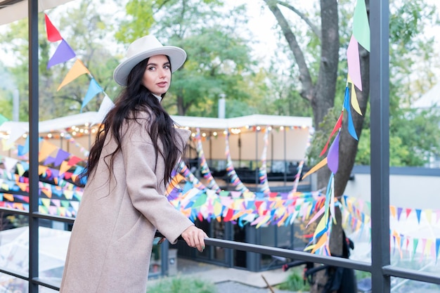 Una bella donna con cappotto e cappello si voltò verso la telecamera, in piedi vicino alla ringhiera al secondo piano. Sullo sfondo di una città decorata con bandiere colorate