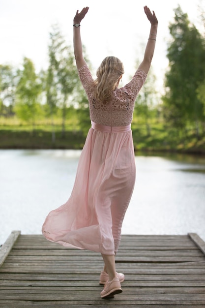 Una bella donna con un vestito rosa fluente si trova su un ponte di legno con le spalle alla telecamera