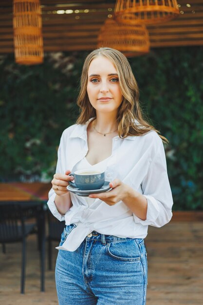 Una bella donna con una camicia bianca e jeans sta bevendo caffè sulla terrazza di un caffè estivo e si sta godendo la sua vacanza