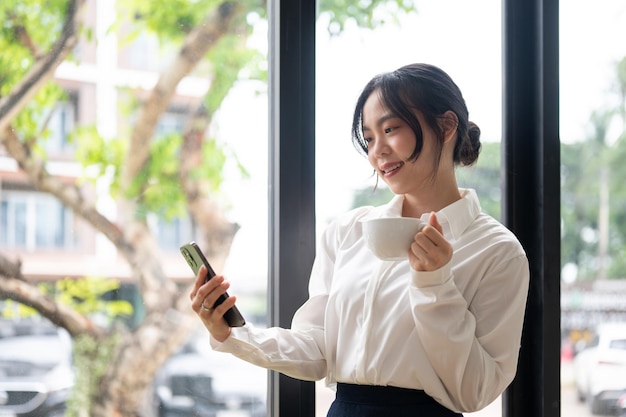 Una bella donna d'affari asiatica che sorseggia caffè e usa il telefono mentre è in piedi vicino alla finestra