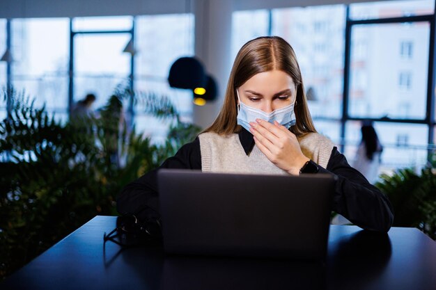 Una bella donna d'affari intelligente in una grande azienda tiene un incontro online con partner commerciali in una maschera protettiva durante il coronavirus si siede a un tavolo in un posto di lavoro con un laptop