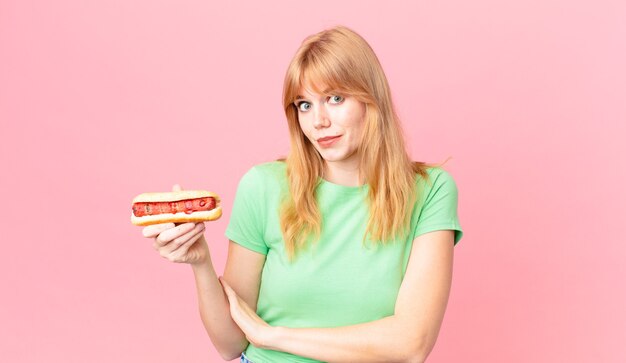 Una bella donna dai capelli rossi che alza le spalle, si sente confusa e incerta e tiene in mano un hot dog