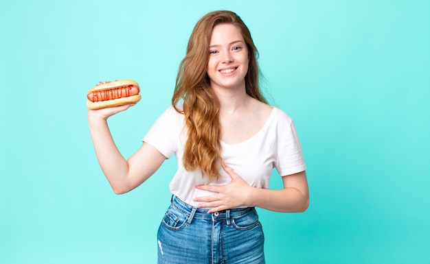 Una bella donna dai capelli rossi che ride ad alta voce per uno scherzo esilarante e tiene in mano un hot dog