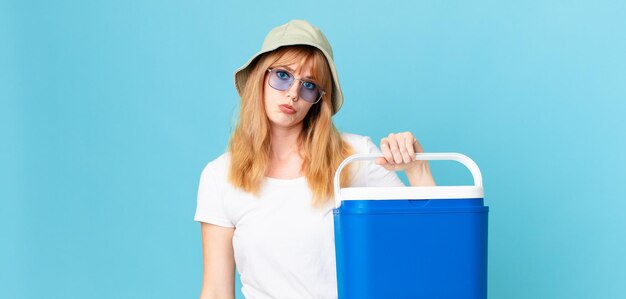 Una bella donna dai capelli rossi che si sente triste e piagnucolona con uno sguardo infelice e piange e tiene in mano un frigorifero portatile. concetto di estate