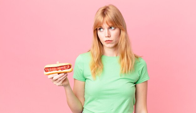 Una bella donna dai capelli rossi che si sente triste, sconvolta o arrabbiata e guarda di lato e tiene in mano un hot dog