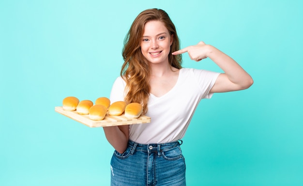 Una bella donna dai capelli rossi che sorride con sicurezza indicando il proprio ampio sorriso e tenendo in mano un panino troy