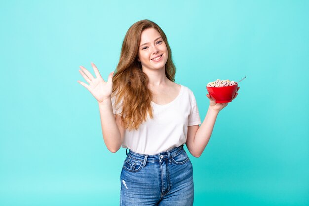 Una bella donna dai capelli rossi che sorride e sembra amichevole, mostra il numero cinque e tiene in mano una ciotola per la colazione