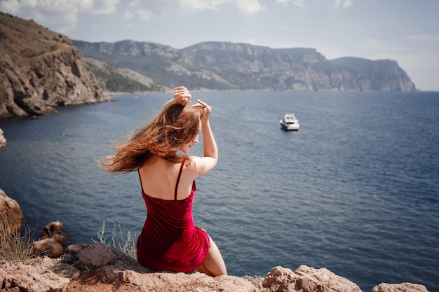 Una bella donna dai capelli rossi in abito rosso è seduta su una roccia con una splendida vista sul paesaggio marino. Pomeriggio estivo godimento di libertà e solitudine
