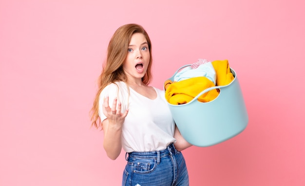 Una bella donna dai capelli rossi stupita, scioccata e stupita con un'incredibile sorpresa e con in mano un cesto di lavaggio con i vestiti