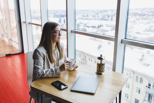 Una bella donna di aspetto sloveno un manager con una giacca leggera e occhiali una ragazza con un sorriso sul viso si siede a un tavolo di legno in un caffè Lavoro a distanza