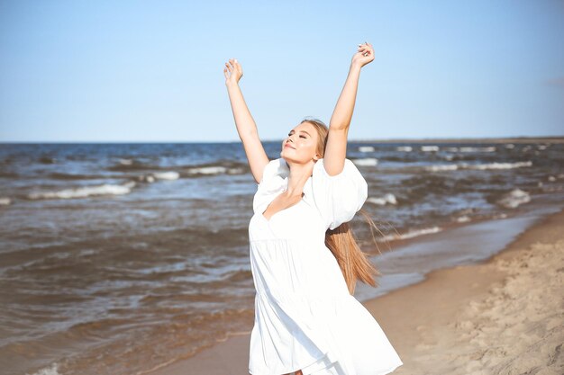 Una bella donna felice e sorridente sulla spiaggia dell'oceano in piedi con un vestito bianco d'estate, alzando le mani.