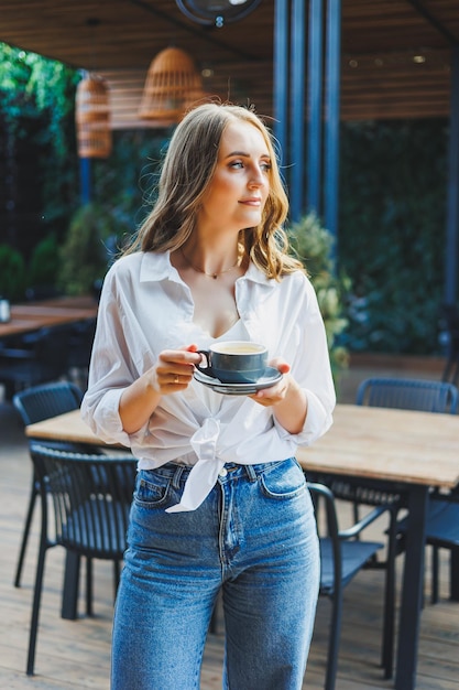 Una bella donna in camicia bianca e jeans sta bevendo caffè sulla terrazza di un bar estivo e si sta godendo la vacanza